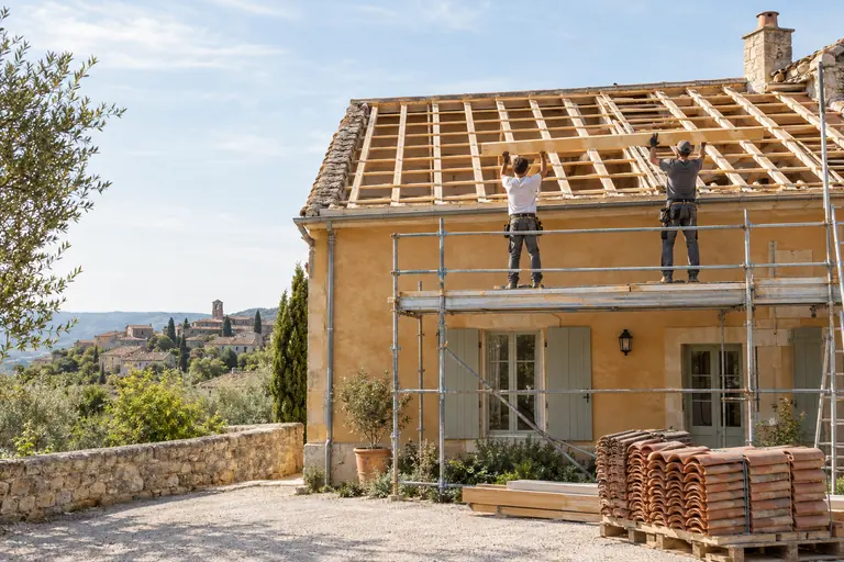 Toiture de maison de village découverte, chevrons et liteaux neufs, deux charpentiers de dos mettant une poutre en place