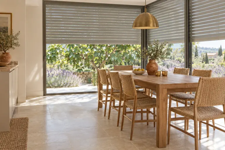 Coin repas dans une extension de cuisine vitrée, table en bois, stores extérieurs à demi baissés et jardin méditerranéen