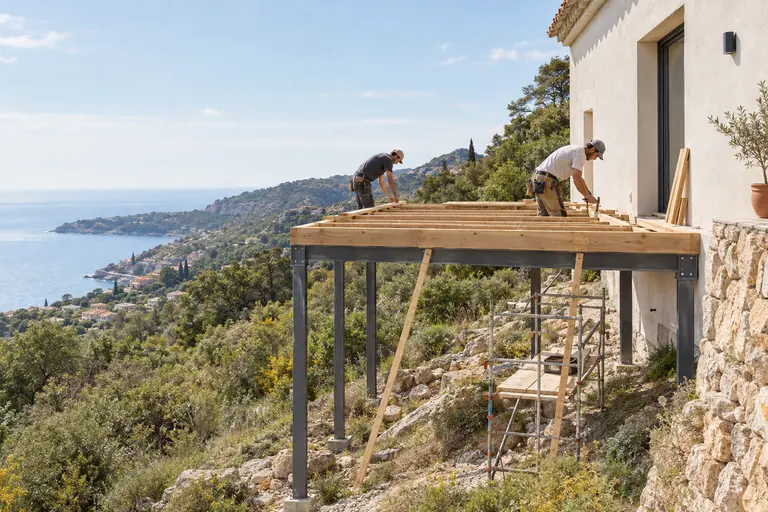 Deux ouvriers de profil assemblant le plancher bois d’une extension posée sur des pilotis en acier sur un terrain en pente