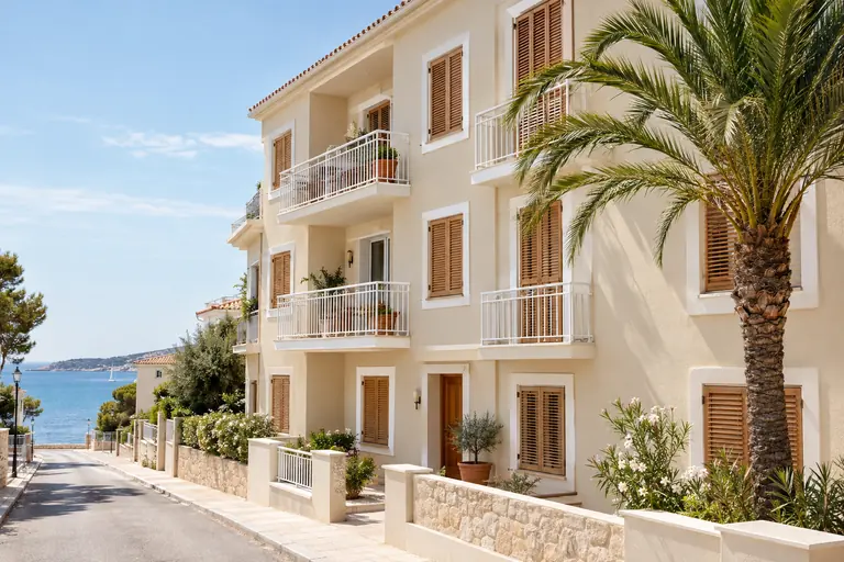 Façade de petit immeuble en bord de mer repeinte en ton sable clair, garde-corps blancs, volets bois et palmier devant l’entrée