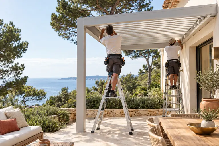 Deux poseurs de dos sur escabeaux montant une pergola bioclimatique en aluminium sur la terrasse d’une villa du Var