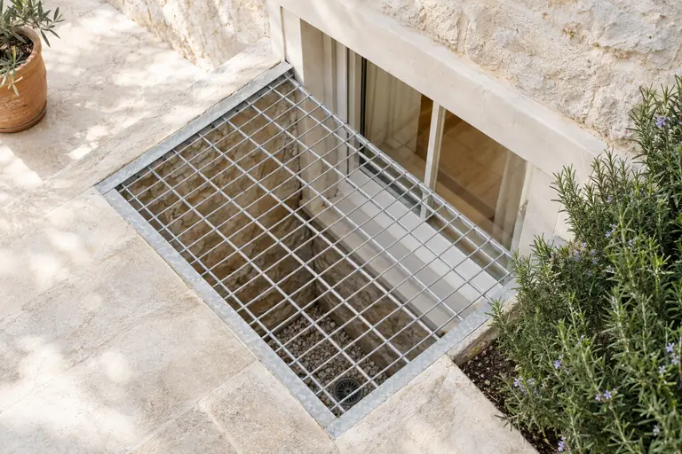 Fosse maçonnée devant une fenêtre de sous-sol, grille galvanisée au-dessus et évacuation d’eau au fond, lumière tombant sur la fenêtre